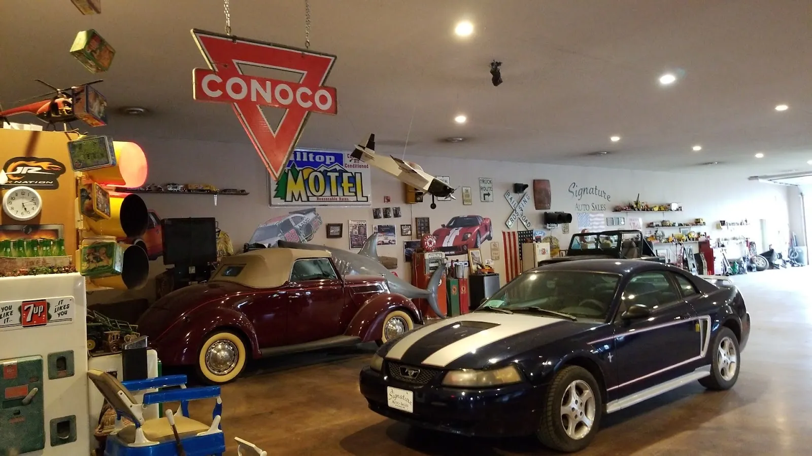 The showroom floor — a dark Mustang and a red Beetle convertible parked indoors beneath a Conoco sign and a Hilltop Motel neon.